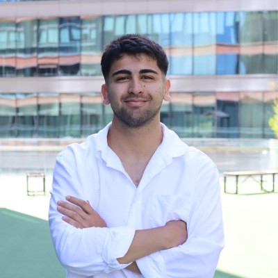 Sefa Kartav, beyaz gömlekle kollarını kavuşturmuş, cam cepheli bir binanın önünde gülümsüyor